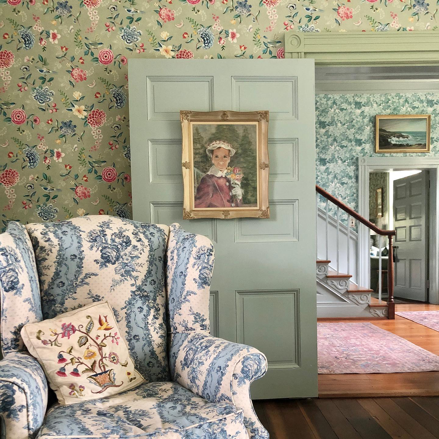 Charming English-inspired drawing room featuring floral wallpaper with a sage green backdrop and pink and blue flowers, a blue and cream floral wingback chair, and a portrait of a lady wearing a bonnet